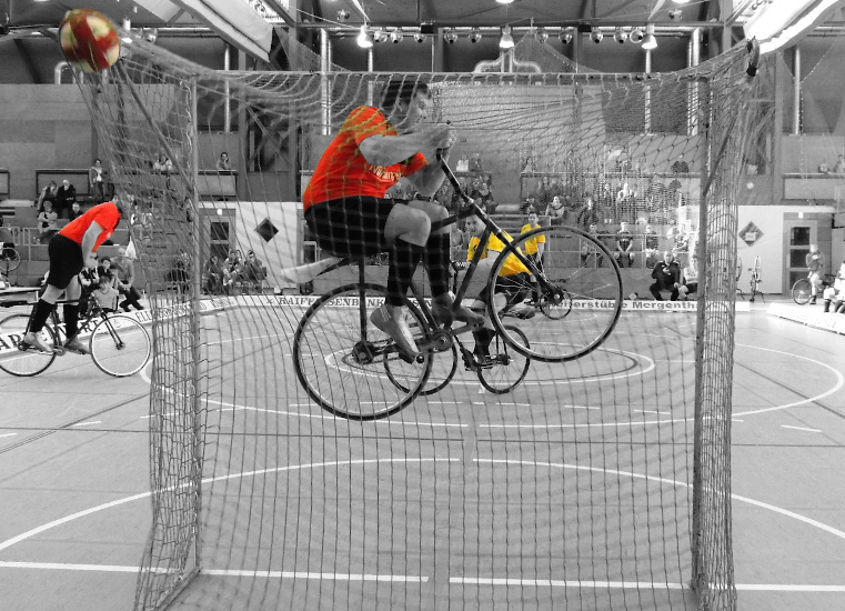Bild eines Radballspiels, wobei der Torwart versucht einen auf das Tor geschossenen Ball zu fangen, indem er mitsamt Fahrrad in die Luft springt.