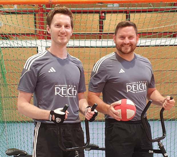 Foto zweier Radballspieler, die dafür mit Radball vor einem Radballtor posieren.