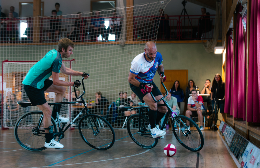 Foto eines Radballspiels, bei dem gerade die beiden Feldspieler dem Ball hinterher jagen.